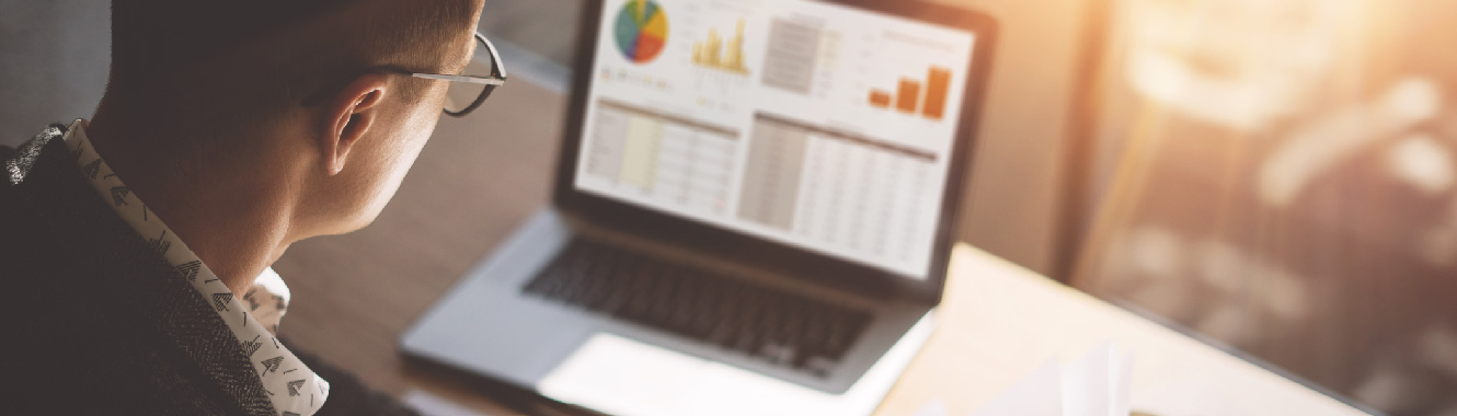 An over-the-shoulder view of a young businessman looking at graphs and charts on a laptop.